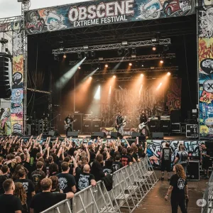 Širokoúhlý záběr pódia na festivalu Obscene Extreme, kde grindcoreová kapela hraje pro rozvášněný dav v mosh pitu pod ostrými světly.